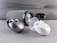 A collection of wireless earbuds is arranged on a flat surface, accompanied by a cylindrical portable speaker in the background. The earbuds vary in color, with one pair being metallic silver and another white, both in stylish, rounded charging cases. The speaker has a fabric exterior with a sleek design.