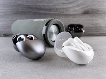 A collection of wireless earbuds is arranged on a flat surface, accompanied by a cylindrical portable speaker in the background. The earbuds vary in color, with one pair being metallic silver and another white, both in stylish, rounded charging cases. The speaker has a fabric exterior with a sleek design.