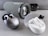 Various wireless earbuds and a portable speaker are arranged on a gray surface. A black and metallic case contains a pair of earbuds, positioned next to a white case with another pair of earbuds. In the background, a cylindrical speaker with a fabric exterior displays its brand logo. The earbuds and speaker are placed in a well-lit setting, highlighting their sleek and modern design.