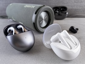 Various wireless earbuds and a portable speaker are arranged on a gray surface. A black and metallic case contains a pair of earbuds, positioned next to a white case with another pair of earbuds. In the background, a cylindrical speaker with a fabric exterior displays its brand logo. The earbuds and speaker are placed in a well-lit setting, highlighting their sleek and modern design.