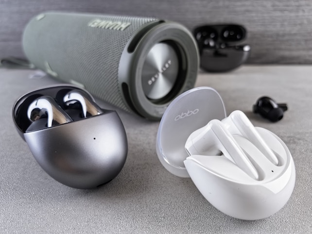 Various wireless earbuds and a portable speaker are arranged on a gray surface. A black and metallic case contains a pair of earbuds, positioned next to a white case with another pair of earbuds. In the background, a cylindrical speaker with a fabric exterior displays its brand logo. The earbuds and speaker are placed in a well-lit setting, highlighting their sleek and modern design.
