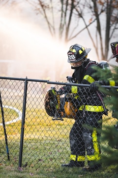 A firefighter wearing a uniform with reflective yellow stripes stands beside a chain-link fence, holding a large power tool. The background is filled with a misty spray, creating a dramatic scene with trees out of focus.