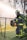 A firefighter wearing a uniform with reflective yellow stripes stands beside a chain-link fence, holding a large power tool. The background is filled with a misty spray, creating a dramatic scene with trees out of focus.