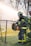 A firefighter wearing a uniform with reflective yellow stripes stands beside a chain-link fence, holding a large power tool. The background is filled with a misty spray, creating a dramatic scene with trees out of focus.