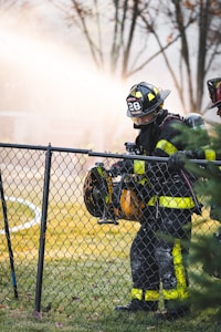 A firefighter wearing a uniform with reflective yellow stripes stands beside a chain-link fence, holding a large power tool. The background is filled with a misty spray, creating a dramatic scene with trees out of focus.