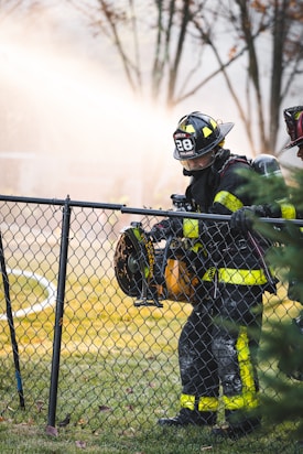 A firefighter wearing a uniform with reflective yellow stripes stands beside a chain-link fence, holding a large power tool. The background is filled with a misty spray, creating a dramatic scene with trees out of focus.