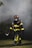 A firefighter, dressed in full gear including a helmet and a protective mask, walks through a smoky environment. The uniform is black with bright yellow reflective strips. The firefighter appears to be carrying a tool, possibly an axe, and the setting suggests an emergency scene with smoke in the air.