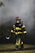 A firefighter, dressed in full gear including a helmet and a protective mask, walks through a smoky environment. The uniform is black with bright yellow reflective strips. The firefighter appears to be carrying a tool, possibly an axe, and the setting suggests an emergency scene with smoke in the air.