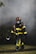 A firefighter, dressed in full gear including a helmet and a protective mask, walks through a smoky environment. The uniform is black with bright yellow reflective strips. The firefighter appears to be carrying a tool, possibly an axe, and the setting suggests an emergency scene with smoke in the air.