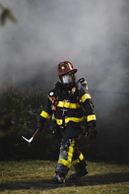 A firefighter, dressed in full gear including a helmet and a protective mask, walks through a smoky environment. The uniform is black with bright yellow reflective strips. The firefighter appears to be carrying a tool, possibly an axe, and the setting suggests an emergency scene with smoke in the air.