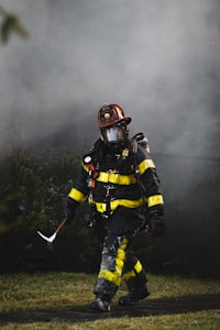 A firefighter, dressed in full gear including a helmet and a protective mask, walks through a smoky environment. The uniform is black with bright yellow reflective strips. The firefighter appears to be carrying a tool, possibly an axe, and the setting suggests an emergency scene with smoke in the air.