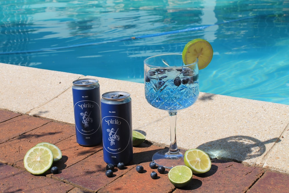 a glass of water and two cans of soda next to a swimming pool