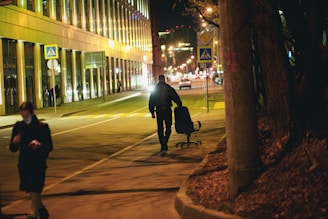 A nighttime urban street scene with a person walking down the sidewalk, pushing a rolling office chair. The area is illuminated by streetlights and lights from surrounding buildings. Another person is walking on the sidewalk looking at a smartphone. Trees and a streetlamp line the side of the street.