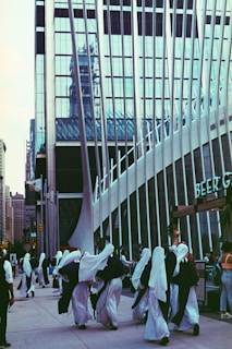 A group of people dressed in white robes and head coverings walk along a city street. Behind them is a modern building with reflective glass panels and vertical metal beams. Additional pedestrians are visible in the background, and the overall scene has an urban and bustling atmosphere.