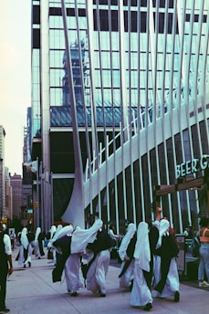 A group of people dressed in white robes and head coverings walk along a city street. Behind them is a modern building with reflective glass panels and vertical metal beams. Additional pedestrians are visible in the background, and the overall scene has an urban and bustling atmosphere.