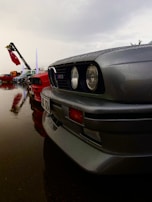 A row of classic cars, including a silver BMW with an M3 badge, is parked on a wet surface with raindrops visible on the vehicles. Behind the line of cars, flags and a crane with a red cloth can be seen, with a cloudy sky overhead.