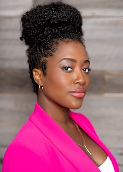 A woman with curly, dark hair styled in an updo, wearing a bright pink blazer over a white top. She has subtle makeup, including eyeliner, eyeshadow, and pink lipstick. A pendant necklace is visible around her neck, and she wears simple hoop earrings. The background consists of wooden paneling.