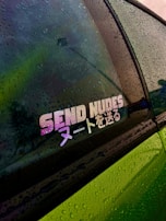 A vehicle's window is covered in raindrops, showcasing a colorful sticker with the phrase 'SEND NUDES' in English and Japanese characters. The window reflects hues of green, purple, and yellow, while the body of the car appears to be green.
