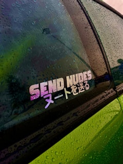 A vehicle's window is covered in raindrops, showcasing a colorful sticker with the phrase 'SEND NUDES' in English and Japanese characters. The window reflects hues of green, purple, and yellow, while the body of the car appears to be green.