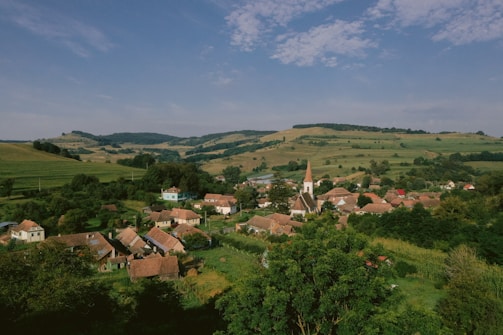 A scenic rural landscape with a village nestled among rolling green hills. The village features a prominent church with a tall steeple, surrounded by houses with red and brown roofs. Lush vegetation and trees are abundant, with a backdrop of hills and a partially cloudy sky.