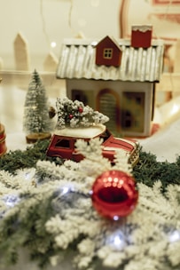 A vintage toy car in red with a snow-covered Christmas tree on its roof sits on a bed of faux snow and green garland. In the background, a miniature house with a snow-dusted roof and small pine trees enhances the wintery holiday scene. A red Christmas ornament and white snowflakes add festive touches.