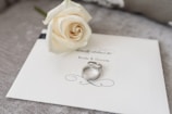A white rose lies delicately on an elegant wedding invitation card labeled 'Bride & Groom.' Two silver rings rest beside the rose, adding a touch of elegance and romance to the setup.