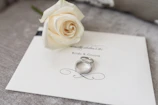 A white rose lies delicately on an elegant wedding invitation card labeled 'Bride & Groom.' Two silver rings rest beside the rose, adding a touch of elegance and romance to the setup.