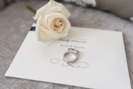 A white rose lies delicately on an elegant wedding invitation card labeled 'Bride & Groom.' Two silver rings rest beside the rose, adding a touch of elegance and romance to the setup.