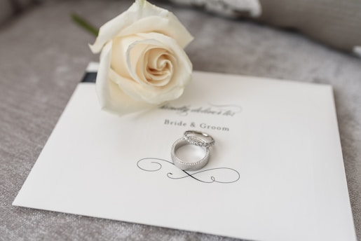 A white rose lies delicately on an elegant wedding invitation card labeled 'Bride & Groom.' Two silver rings rest beside the rose, adding a touch of elegance and romance to the setup.