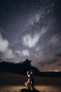 Under a starry night sky, a couple stands lovingly embraced on a sandy terrain illuminated by a soft, warm light. Wispy clouds are scattered across the sky, adding depth to the scene.
