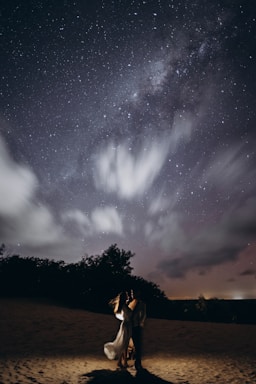 Under a starry night sky, a couple stands lovingly embraced on a sandy terrain illuminated by a soft, warm light. Wispy clouds are scattered across the sky, adding depth to the scene.