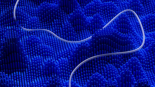 A dynamic scene featuring wavy rows of vertical blue cylinders creating an undulating, textured landscape. A curved, thin, translucent line flows smoothly over the surface, adding contrast and fluidity.