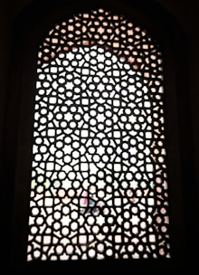 A detailed lattice pattern with geometric shapes is carved into a window screen, allowing blurred light to filter through. The structure consists of repeating star and hexagonal shapes arranged in an intricate design, creating a symmetrical and decorative effect.