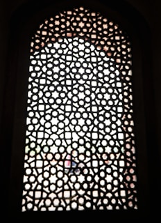 A detailed lattice pattern with geometric shapes is carved into a window screen, allowing blurred light to filter through. The structure consists of repeating star and hexagonal shapes arranged in an intricate design, creating a symmetrical and decorative effect.