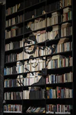 A large bookshelf filled with books of various sizes and colors, organized neatly on multiple shelves. The books themselves form the outline of a face, creating an artistic visual effect. The shelves are predominantly dark, emphasizing the lighter tones of the books.