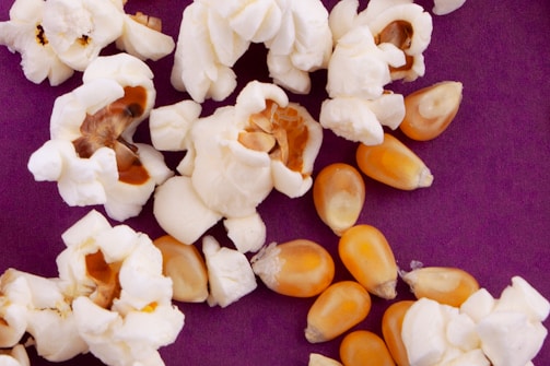A collection of popped popcorn scattered alongside unpopped corn kernels, all set against a rich purple background. The popcorn is fluffy and white, while the kernels are smooth and have a bright golden-yellow hue.