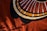A textile design featuring bold geometric patterns with concentric circles and radial lines. The background is a rich, warm red fabric, and shadows of objects like leaves or branches are cast across the surface, adding depth to the composition.