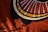 A textile design featuring bold geometric patterns with concentric circles and radial lines. The background is a rich, warm red fabric, and shadows of objects like leaves or branches are cast across the surface, adding depth to the composition.