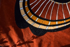 A textile design featuring bold geometric patterns with concentric circles and radial lines. The background is a rich, warm red fabric, and shadows of objects like leaves or branches are cast across the surface, adding depth to the composition.