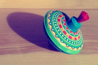 A colorful spinning top with intricate geometric patterns rests on a wooden surface, casting a soft shadow. The top features a combination of red, green, and yellow designs, giving it a vibrant appearance.