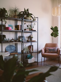 A cozy, well-decorated living space with a metal bookshelf filled with various plants, books, and decorative items. In the corner, a pink armchair with a patterned cushion sits next to a medium-sized plant. The room features a modern light fixture and a striped rug on a wooden floor.