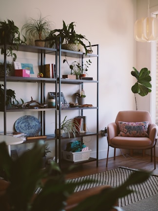 A cozy, well-decorated living space with a metal bookshelf filled with various plants, books, and decorative items. In the corner, a pink armchair with a patterned cushion sits next to a medium-sized plant. The room features a modern light fixture and a striped rug on a wooden floor.