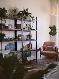 A cozy, well-decorated living space with a metal bookshelf filled with various plants, books, and decorative items. In the corner, a pink armchair with a patterned cushion sits next to a medium-sized plant. The room features a modern light fixture and a striped rug on a wooden floor.