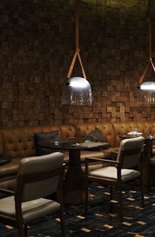 A cozy and elegant restaurant interior with wooden paneled walls and pendant lights hanging above circular tables. The seating arrangement includes padded leather benches and cushioned chairs. The mood is inviting and intimate, with dark, warm colors and soft lighting.