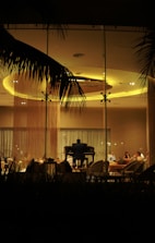 A dimly lit room viewed through a glass window, with a pianist playing a grand piano in the center. Several people are seated around the pianist on comfortable chairs, engaged in conversation. Large palm leaves can be seen framing the scene from above, hinting at an outdoor atmosphere.