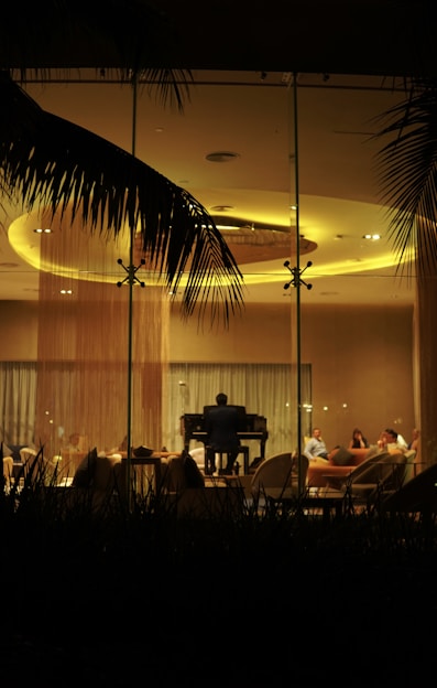 A dimly lit room viewed through a glass window, with a pianist playing a grand piano in the center. Several people are seated around the pianist on comfortable chairs, engaged in conversation. Large palm leaves can be seen framing the scene from above, hinting at an outdoor atmosphere.