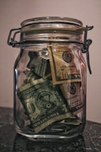 A glass jar filled with various denominations of US dollar bills, including one and ten dollar notes. The jar is placed on a dark surface and is sealed with a metal clasp. The jar appears to be full of folded bills.