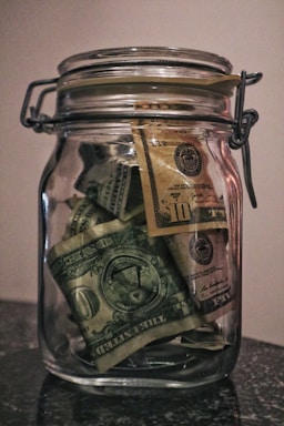 A glass jar filled with various denominations of US dollar bills, including one and ten dollar notes. The jar is placed on a dark surface and is sealed with a metal clasp. The jar appears to be full of folded bills.