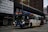 A city bus in motion on a street lined with tall buildings. The bus is marked with 'Rea Vaya' branding and is predominantly white with blue and red accents. The surrounding area features signage advertising flat rentals, and the street is relatively empty.
