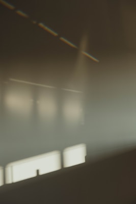 The image features a blurred interior scene with soft lighting and muted colors, creating an abstract and minimalistic atmosphere. There are hints of structural elements such as walls or windows, and the light appears to be diffused, casting subtle reflections.